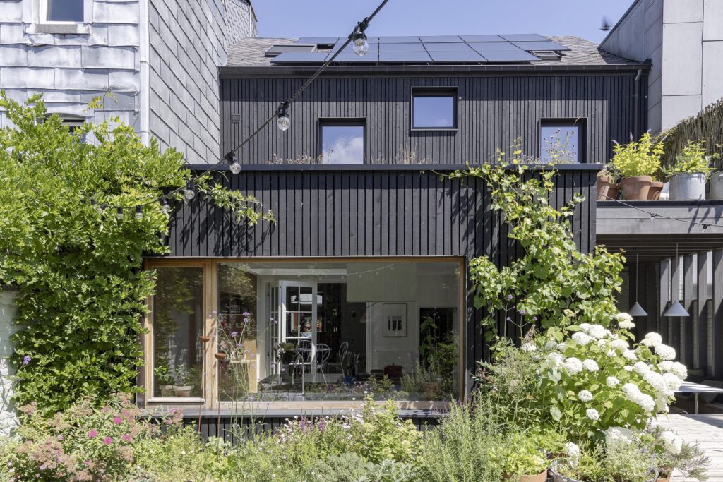 Façade arrière moderne en bois noir avec grandes baies vitrées et jardin fleuri.
