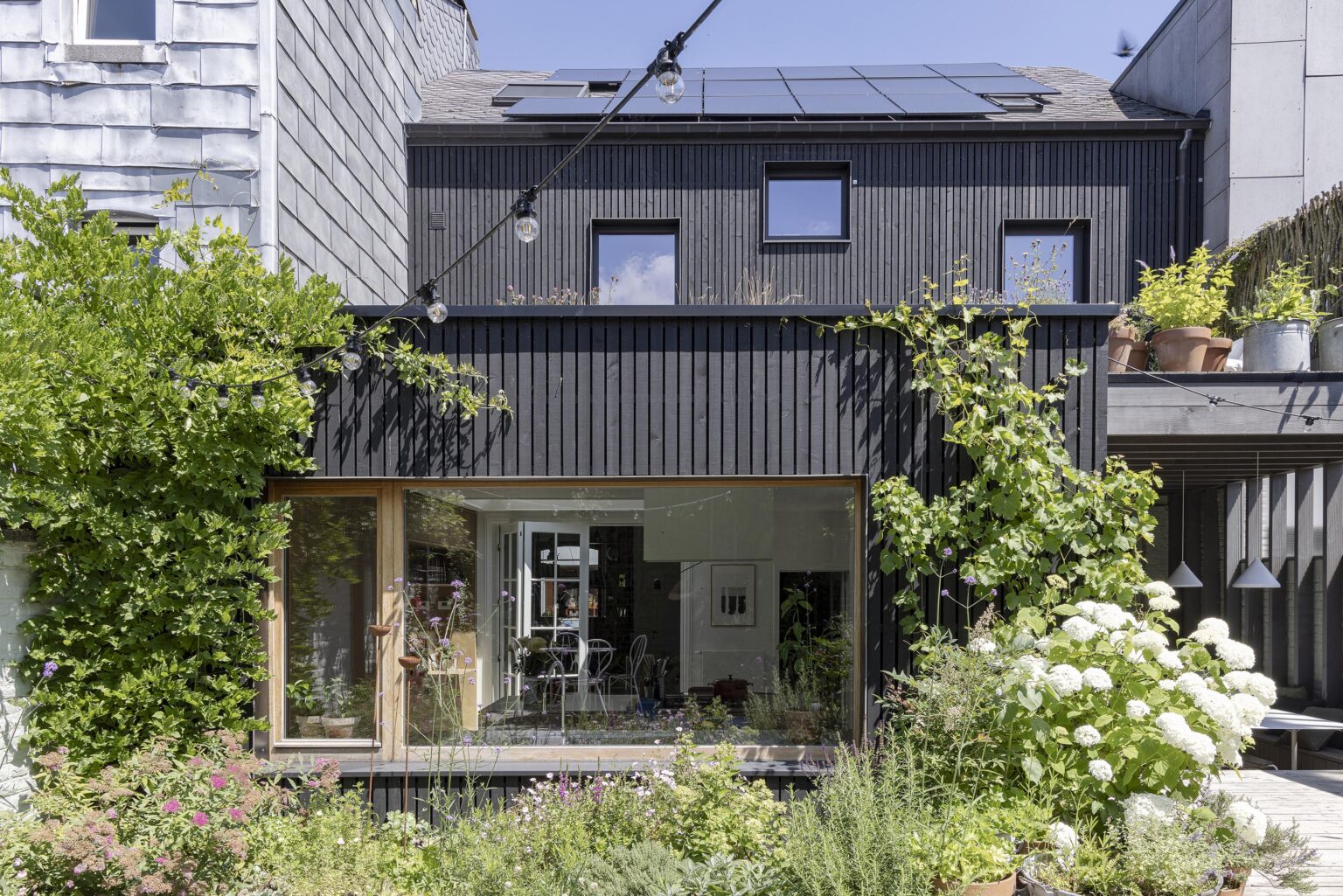 Façade arrière moderne en bois noir avec grandes baies vitrées et jardin fleuri.