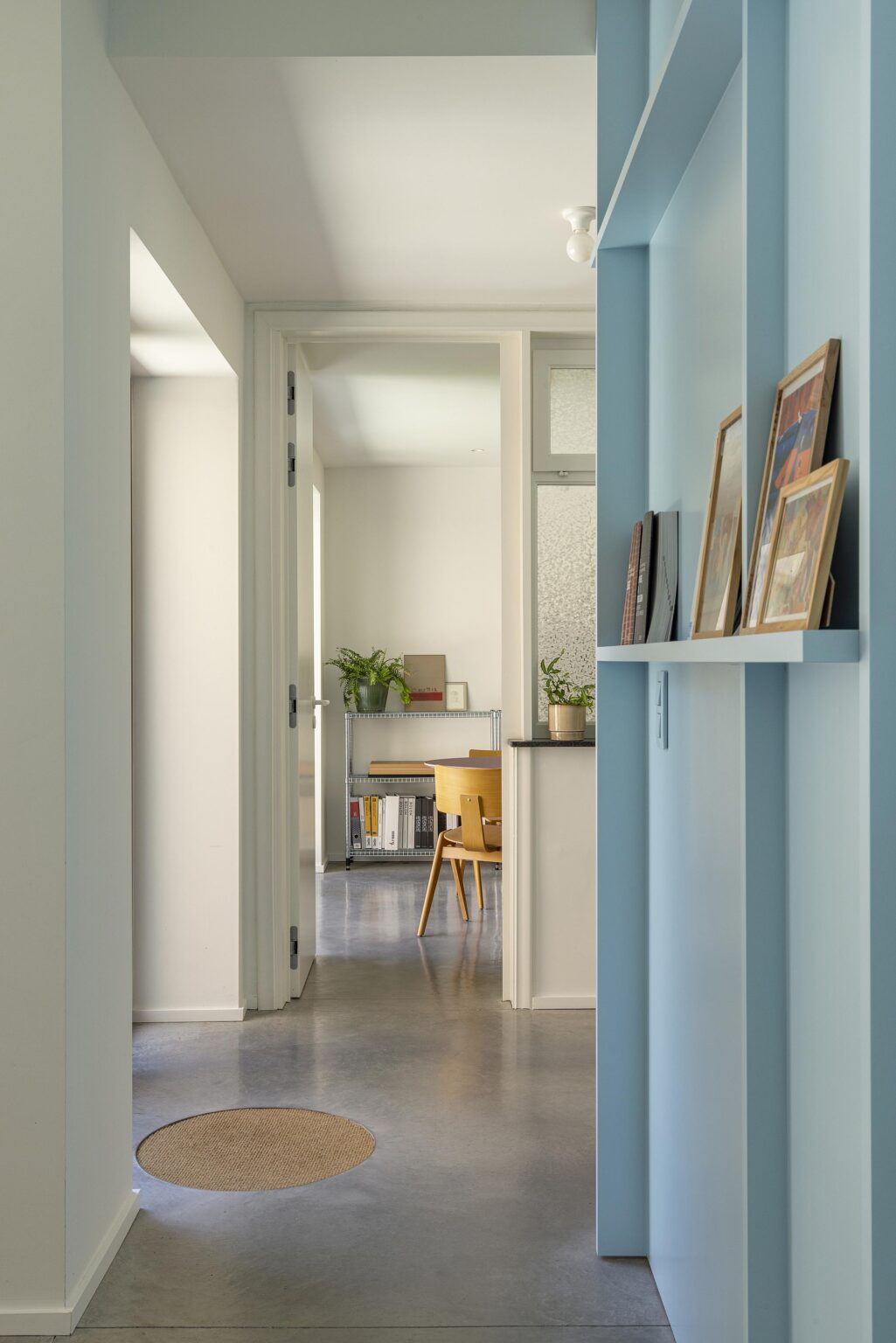 Couloir lumineux avec mur bleu, cadres posés sur une étagère et vue sur une salle avec table et plantes.