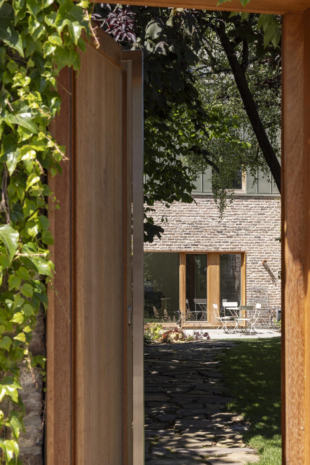 Vue depuis un portail en bois ouvert donnant sur un jardin et une terrasse avec table et chaises blanches.